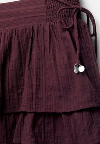 Bordeaux gelaagde rok gemaakt van textuurstof, met een geschuurd tailleband met een trekkoord en decoratieve metalen accenten.