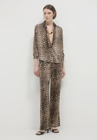 Donna che indossa un blusa coordinata in stampa leopardata e pantaloni a gamba larga con tacchi open-toe neri e collana di perline spesse contro un muro bianco.