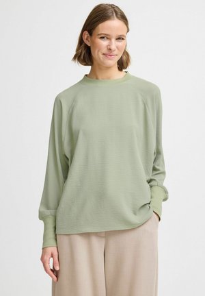 Top lung cu mânecă lungă, de culoare verde deschis, cu guler cu textură ribbed și croială lejeră. Textură moale, cu mâneci raglan. Asortat cu pantaloni bej.