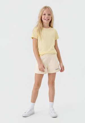 Uśmiechnięte dziecko stojące, ubrane w bladożółtą koszulkę, kremowe szorty z napisem na nodze, białe skarpetki i białe trampki na jednolitym tle.