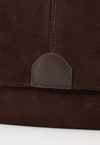Brown suede bag with a textured leather accent. Features a rounded tab detail and contrasting stitching along the edge.
