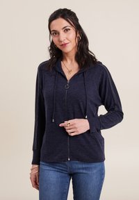 Felpa con zip blu marina, con cordini e dettagli metallici lungo le maniche; texture morbida e vestibilità comoda; indossata con jeans blu.