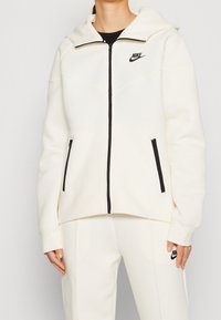 Bílá zipová mikina Nike s vysokým límcem, černým zipem a dvěma bočními zipovými kapsami. Obsahuje prošívané detaily a logo na hrudi.