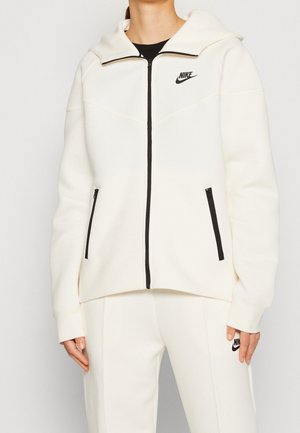 Felpa bianca Nike con zip e colletto alto, zip nera e due tasche laterali con zip. Presenta dettagli cuciti e logo sul petto.