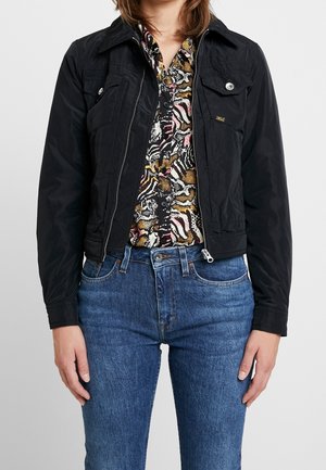 Kobieta ubrana w czarną kurtkę zapinaną na zamek błyskawiczny, na wzorzystej koszuli na guziki i niebieskie, wysokie jeansy, stojąca na jednolitym tle.