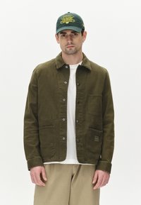 Jachetă verde măsliniu, din material din bumbac, cu închidere butonată, două buzunare frontale, asociată cu o cămașă albă și pantaloni bej deschis, design simplu.