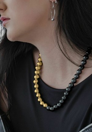 Nahaufnahme einer Person, die eine gold- und schwarzperlige Halskette und einen silbernen, abstrakt geformten Ohrring trägt, mit schwarzem Haar und schwarzer Kleidung.