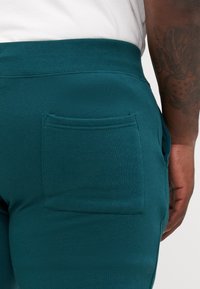 Teal fleece joggers met een geribde tailleband en een patchzak aan de rechterkant, zachte textuur zichtbaar, close-up van de achterkant.