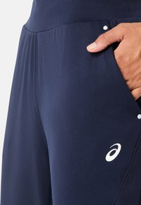 Marine sportieve broeken hebben een gladde, rekbare stof met een zijzak. Een Asics-logo in het wit verschijnt op het onderbeen.