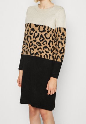 Femme portant une robe en tricot à manches longues avec un haut crème, un milieu à imprimé léopard et une jupe noire unie, posant devant un fond blanc.