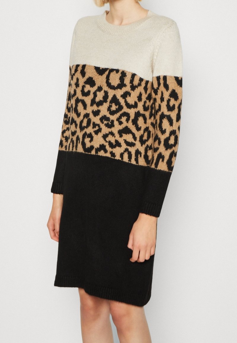 Femme portant une robe en tricot à manches longues avec un haut crème, un milieu à imprimé léopard et une jupe noire unie, posant devant un fond blanc.