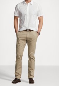 Camisa blanca de manga corta con un logo azul, combinada con pantalones beige y mocasines marrones. Presenta un cuello tipo button-down y un cinturón.