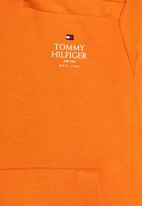 Sudadera naranja con capucha, que cuenta con un bolsillo canguro en la parte frontal, textura suave y un logotipo bordado en blanco con una pequeña bandera tricolor encima.