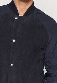 Chaqueta bomber azul marino con cuerpo de ante, mangas de nylon, cuello con elástico acanalado y botones a presión plateados. Textura suave y diseño limpio y moderno.