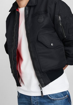 Blouson bomber noir zippé avec poche à rabat et fermeture éclair au bras, porté ouvert sur une chemise blanche et un jean bleu, sur une personne du torse aux hanches.