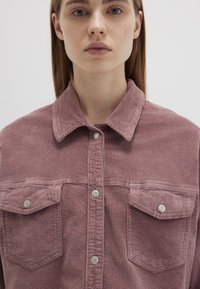 Cordhemd in sanftem Mauve mit Kragen, zwei Brusttaschen und silbernen Druckknöpfen. Es hat eine gerippte Textur und einen lässigen Schnitt.