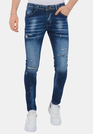 Slim-fit blauwe distressed jeans met vervaagde whiskering en verfspatdetails, gedragen met witte sneakers en zilveren armbanden.