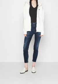 Vit zip-up hoodie, svart v-ringad t-shirt, mörkblå skinny jeans med ljus slitning och vita slip-on skor. Enkelt design och avslappnad passform.