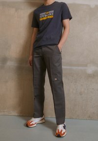 Personne portant un t-shirt noir New Balance, un pantalon Dickies gris foncé et des baskets orange et blanches, debout contre un mur en béton beige.