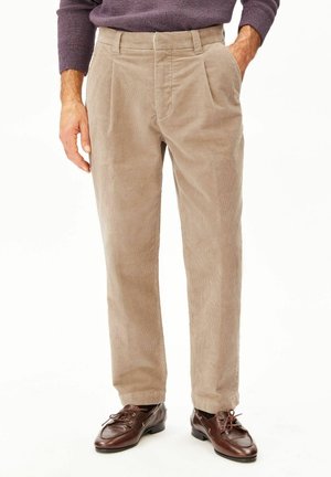 Mann trägt beige Cordhosen mit Vorderfalten, kombiniert mit dunkelbraunen Leder-Schnürschuhen und einem lila Pullover.