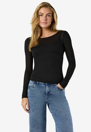 Giovane donna con lunghi capelli biondi che indossa una maglietta nera aderente a maniche lunghe e jeans blu, con una mano in tasca, su sfondo bianco.