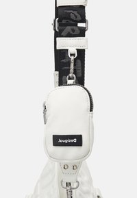 Mini pochette zippée blanche avec logo Desigual noir, attachée à une sangle noire ajustable avec des accents en cuir blanc.
