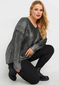Schwarz und silber gestreifter Strickpullover mit V-Ausschnitt und lockerer Passform, getragen über schwarzen Hosen und Stiefeletten. Verfügt über einen strukturierten Stoff und lange Ärmel.