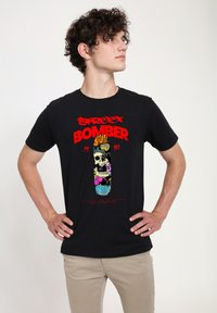 Camiseta negra con un diseño gráfico colorido que presenta una calavera, flores y el texto "BOMBER 1990". De mangas cortas y estilo de cuello redondo.