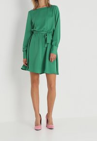 Grünes langärmliges Kleid mit Taillengürtel, aus glattem Stoff gefertigt. Verfügt über einen runden Ausschnitt und einen ausgestellten Rock. Kombiniert mit pinken High Heels.