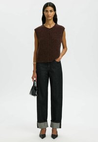 Brązowy bezrękawnik dzianinowy z okrągłym dekoltem i czarnym zapięciem na guziki, zestawiony z ciemnymi dżinsami z wysokim stanem z podwiniętymi szarymi mankietami.