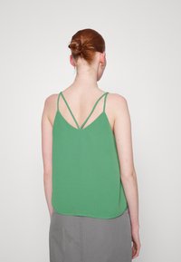 Žena s červenými vlasy v drdolu, oblečená ve zeleném tílku s ramínky a šedé sukni, zobrazená zezadu na neutrálním pozadí.