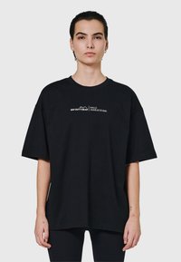 Ung person med kort mörkt hår bär en oversized svart T-shirt med liten vit text "OFF DUTY GEAR HALO" på bröstet mot en enkel ljus bakgrund.