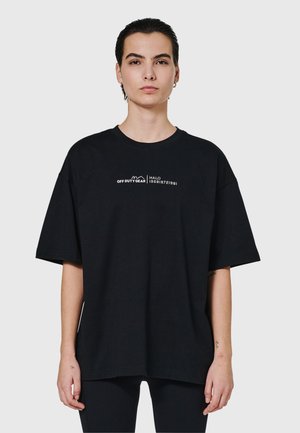 Jong persoon met kort donker haar draagt een oversized zwart T-shirt met kleine witte tekst "OFF DUTY GEAR HALO" op de borst tegen een effen lichte achtergrond.