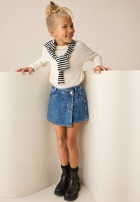 Denimrok met een onafgewerkte zoom en knopen aan de voorkant, gecombineerd met een crèmekleurig shirt met lange mouwen en een sjaal met zwarte strepen, afgemaakt met zwarte laarzen.