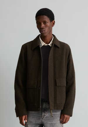Junger Mann trägt eine dunkelbraune Wolljacke über einem schwarzen Pullover und Hemd mit Kragen, steht vor einem schlichten hellgrauen Hintergrund.
