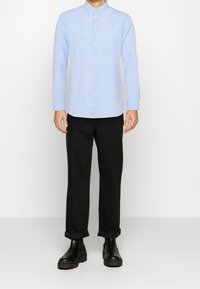 Camicia azzurro chiaro con colletto, maniche lunghe e bottoni frontali, abbinata a pantaloni neri arrotolati e stivaletti neri.