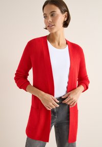 Rote Strickjacke mit langen Ärmeln, offener Vorderseite und aufgesetzten Taschen. Getragen über einem weißen Hemd und kombiniert mit grauen Jeans.