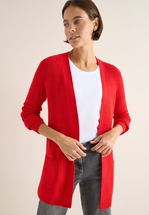 Cardigan rosso lavorato a maglia con maniche lunghe, frontale aperto e tasche a toppa. Indossato su una camicia bianca e abbinato a jeans grigi.