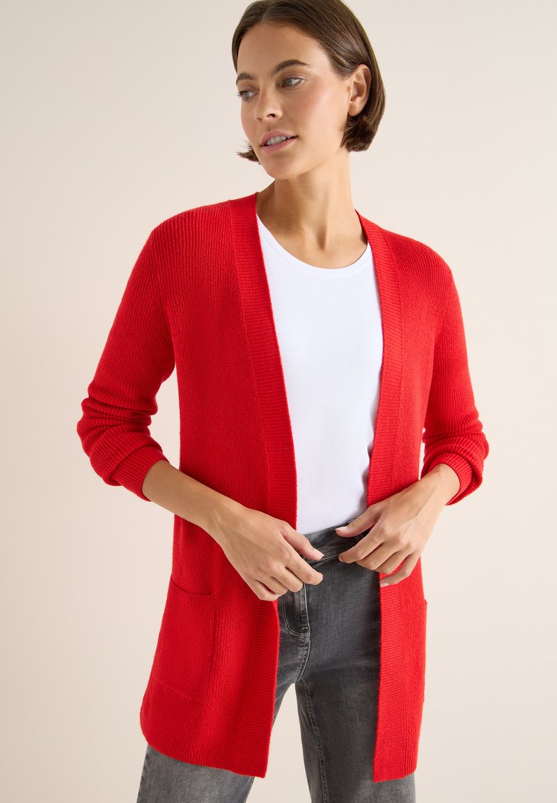 Rote Strickjacke mit langen Ärmeln, offener Vorderseite und aufgesetzten Taschen. Getragen über einem weißen Hemd und kombiniert mit grauen Jeans.