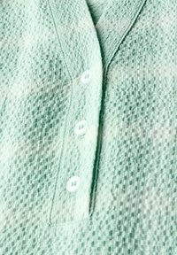 Camicia button-up verde chiaro realizzata in un tessuto testurizzato con piccoli motivi tessuti e tre bottoni bianchi lungo la pattina anteriore.