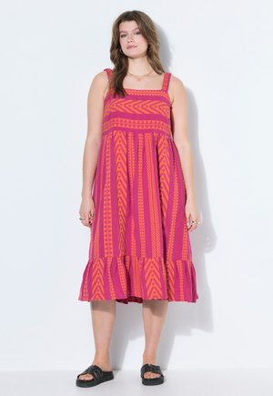 Kvinde iført en ærmeløs midi-kjole i pink og orange med mønster og flæsekant samt sorte slippers, stående mod en hvid baggrund.