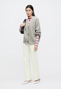 Šedý pletený kardigan s výstřihem do V a zapínáním na knoflíky, v kombinaci s červeným pruhovaným topem s dlouhým rukávem, krémovými kalhotami a bílými špičatými botami.