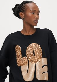 Sweatshirt preta com gráfico "LOVE" em dourado e bronze cintilantes. Textura macia, corte descontraído, com gola e punhos canelados.