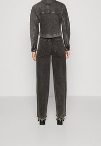 Osoba oblečená v černé džínové bundě s efektem acid wash a džíny s vysokým pasem a širokými nohavicemi, stojící čelem k jednobarevnému světle šedému pozadí.