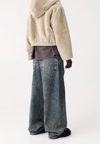 Beige faux fur hættejakke, lagdelt over en grå skjorte, parret med brede blå denimjeans med et vertikalt stribemønster. Sorte fodtøj.