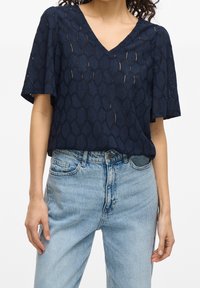 Woman wearing a navy blue short-sleeve V-neck top with a geometric pattern and light blue high-waisted jeans with button and zipper fastening.