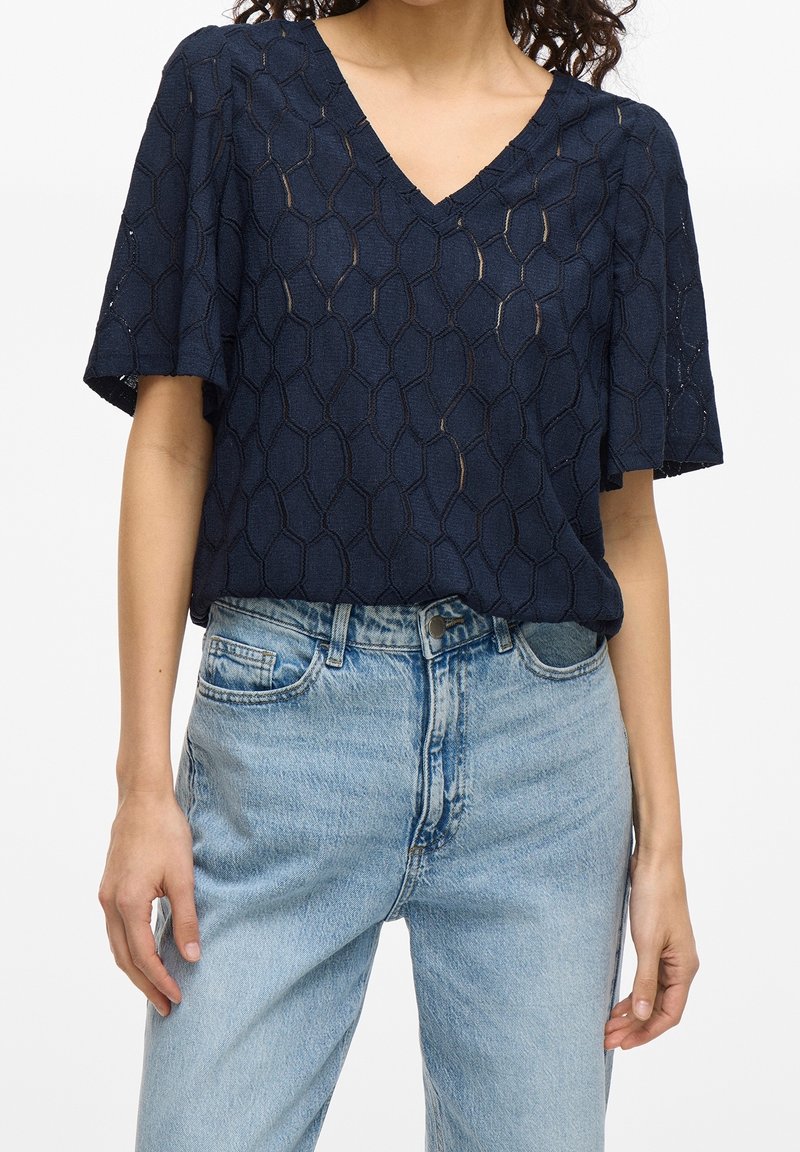 Woman wearing a navy blue short-sleeve V-neck top with a geometric pattern and light blue high-waisted jeans with button and zipper fastening.