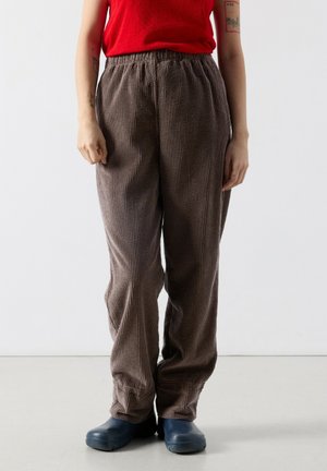 Osoba nosiaca voľné hnedé kordové nohavice, námornícky modré slip-on topánky a červený tielkový top, stojaca na svetlej podlahe s bielym pozadím.