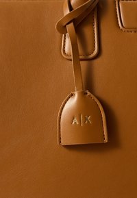 Hnědá kožená kabelka s texturovaným povrchem, obsahující zlatou embosovanou štítek s nápisem "A|X." Obsahuje detail s uzlovým popruhem.