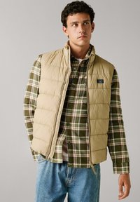 Gilet beige imbottito sopra una camicia a quadri verde con accenti bianchi e rossi. I jeans sono di un blu chiaro. Outfit casual a strati con chiusura a zip.